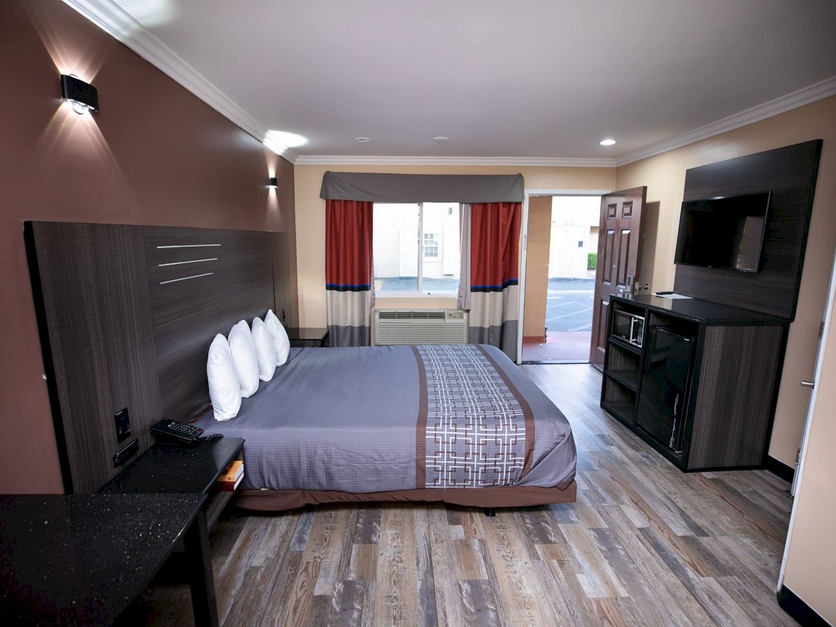 The image shows a hotel room with a bed, TV, microwave, mini-fridge, desk, and window with curtains. The room has modern decor and wooden flooring.