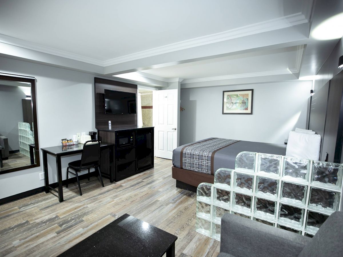 A modern hotel room with a bed, desk, TV, mirror, and sofa. There's a glass partition and a door leading to another area. The flooring is wood.