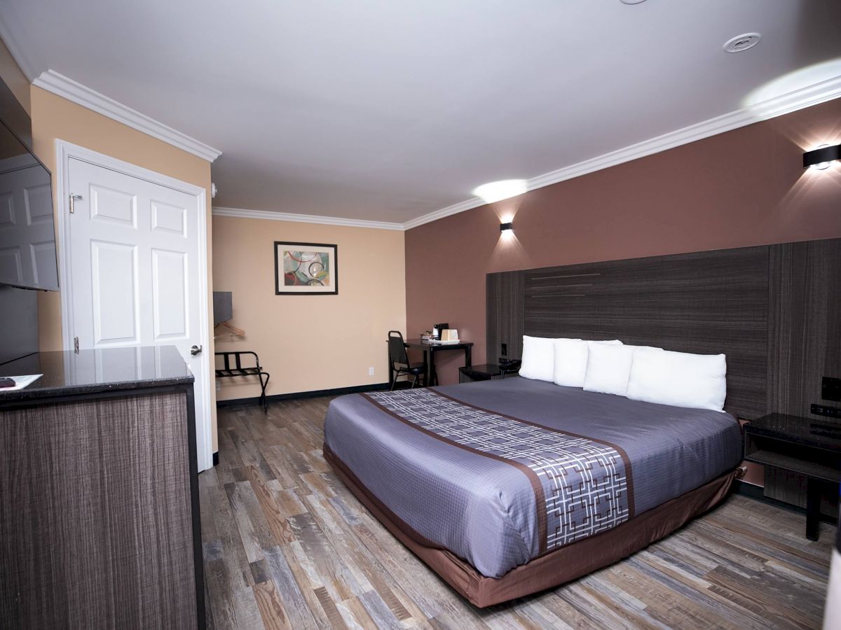 The image shows a modern hotel room with a large bed, white pillows, a desk with a chair, and a TV on a cabinet, and framed artwork on the wall.