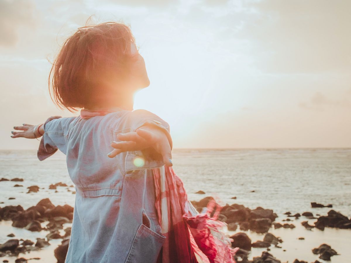 A person stands with outstretched arms facing the sunset by a rocky beach, basking in the warm light and embracing the serene moment.