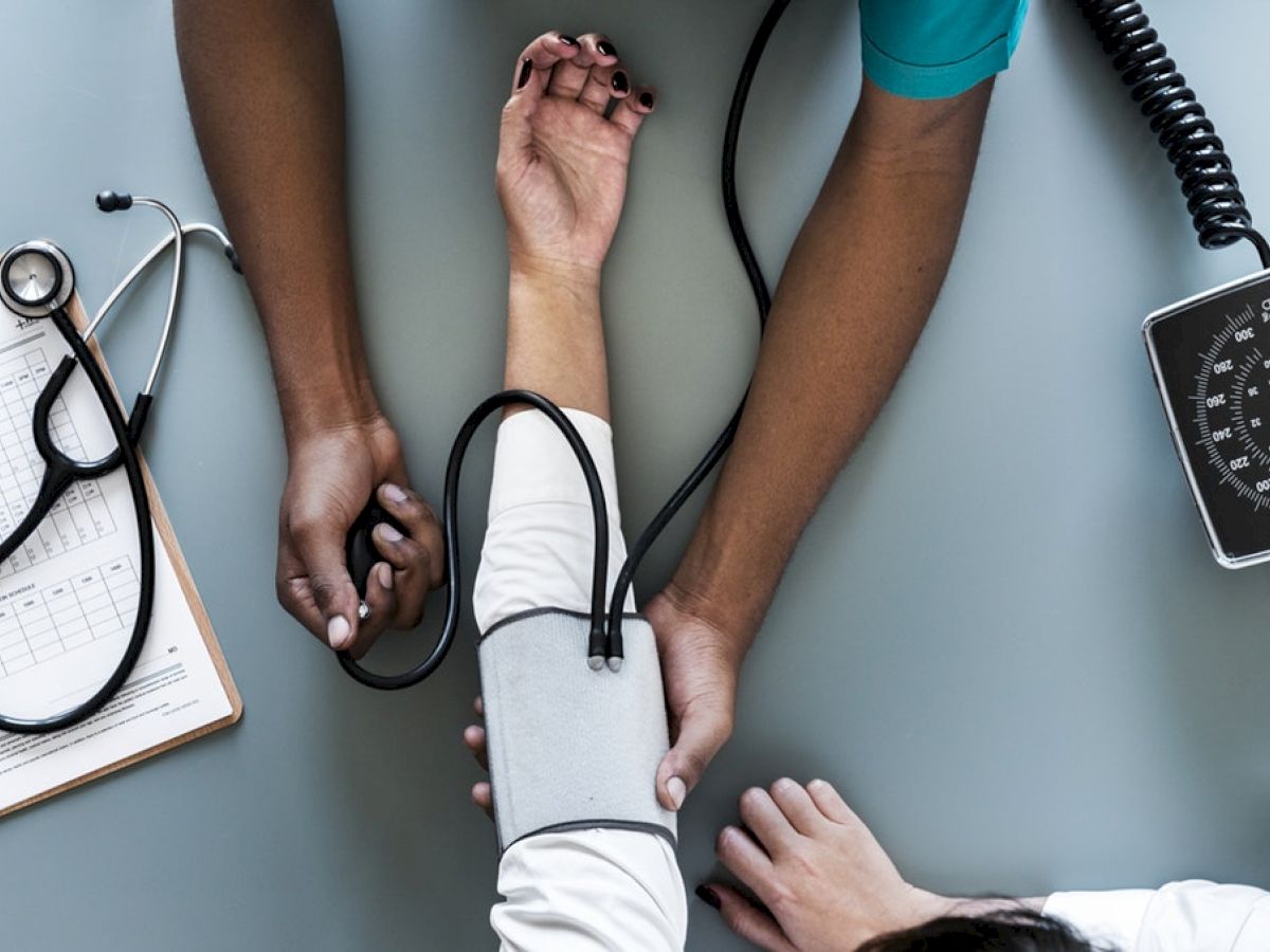 A medical professional is taking a patient's blood pressure using a sphygmomanometer, with a stethoscope and clipboard nearby on the table.