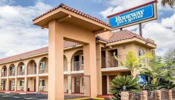 The image shows a tan-colored Rodeway Inn & Suites hotel building with a tiled roof and balconies, surrounded by plants and a clear sky.