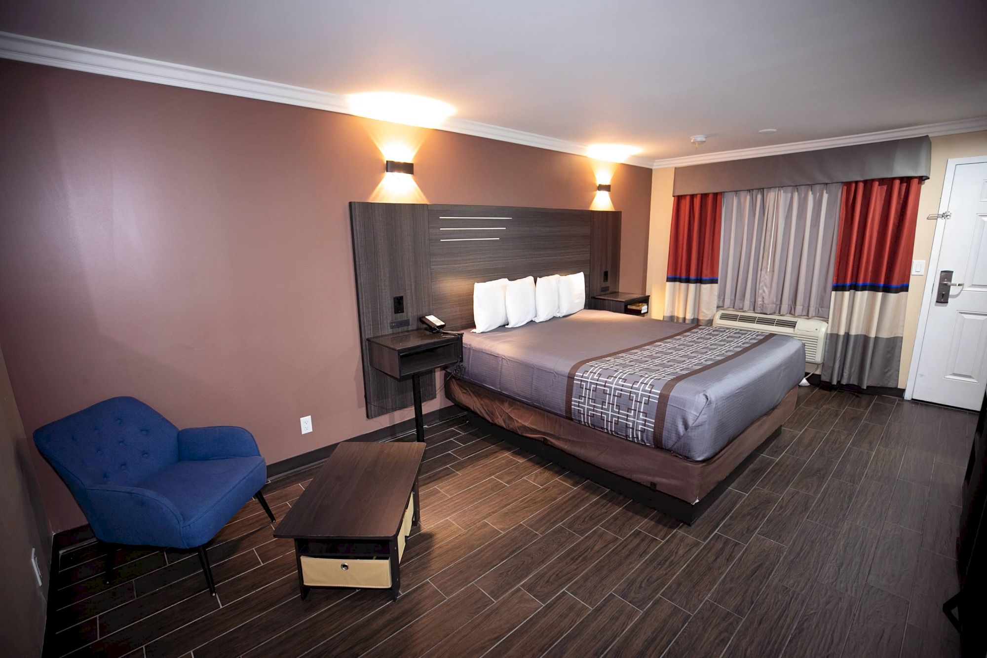 This image shows a modern hotel room with a bed, bedside tables, a blue chair, a small table, and a window with curtains, all under warm lighting.