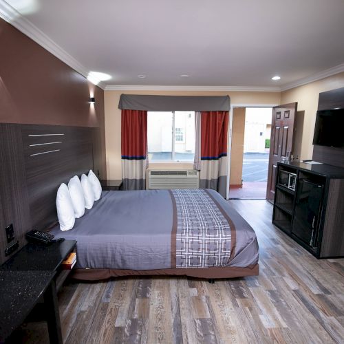 A modern hotel room with a bed, wall-mounted TV, microwave, mini fridge, desk, and a window with red and gray curtains.