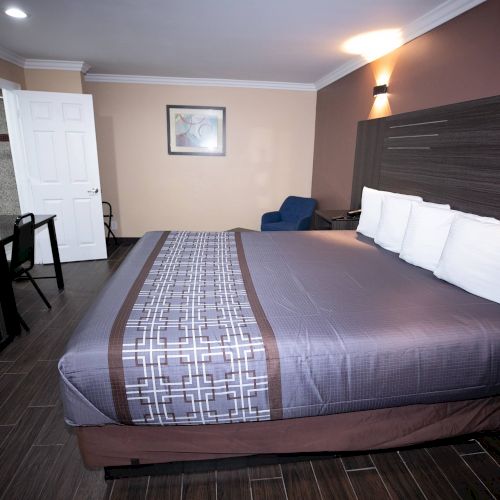 A neatly arranged hotel room with a large bed, white pillows, and a patterned bedspread, featuring a desk, chair, open door, and framed wall art.