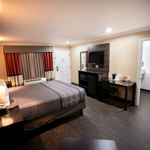 A well-lit hotel room with a large bed, TV, desk, and mini-fridge. Door to a bathroom on the right, and window with curtains on the far wall.