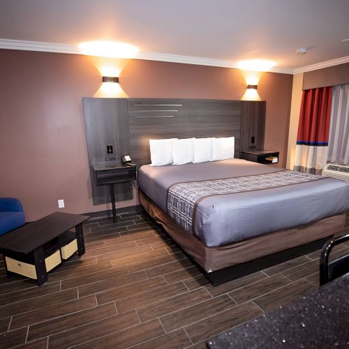 This image shows a hotel room with a king-size bed, a blue chair, a small table, and a window with curtains. The floor is wooden.