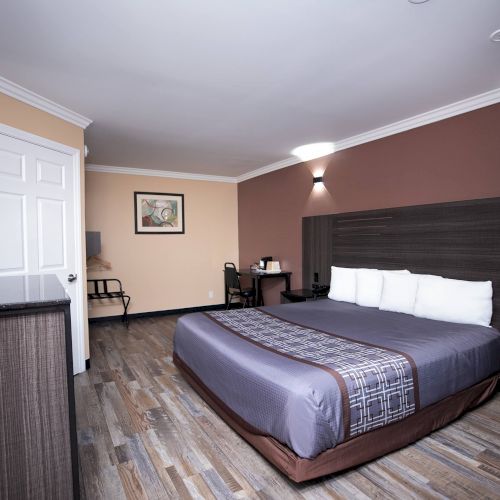 This image shows a hotel room with a large bed, a desk and chair, a TV on a dresser, and modern decor with a wood floor and wall lights.