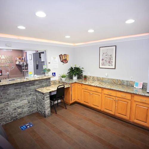 A hotel lobby with a reception desk, a granite countertop, wooden cabinets, a TV, a plant, and a sign that reads 