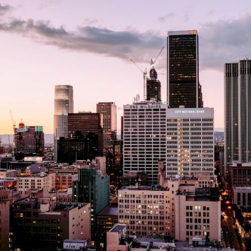 The image shows a city skyline at dusk with various high-rise buildings, construction cranes, and lit windows, under a partially cloudy sky.